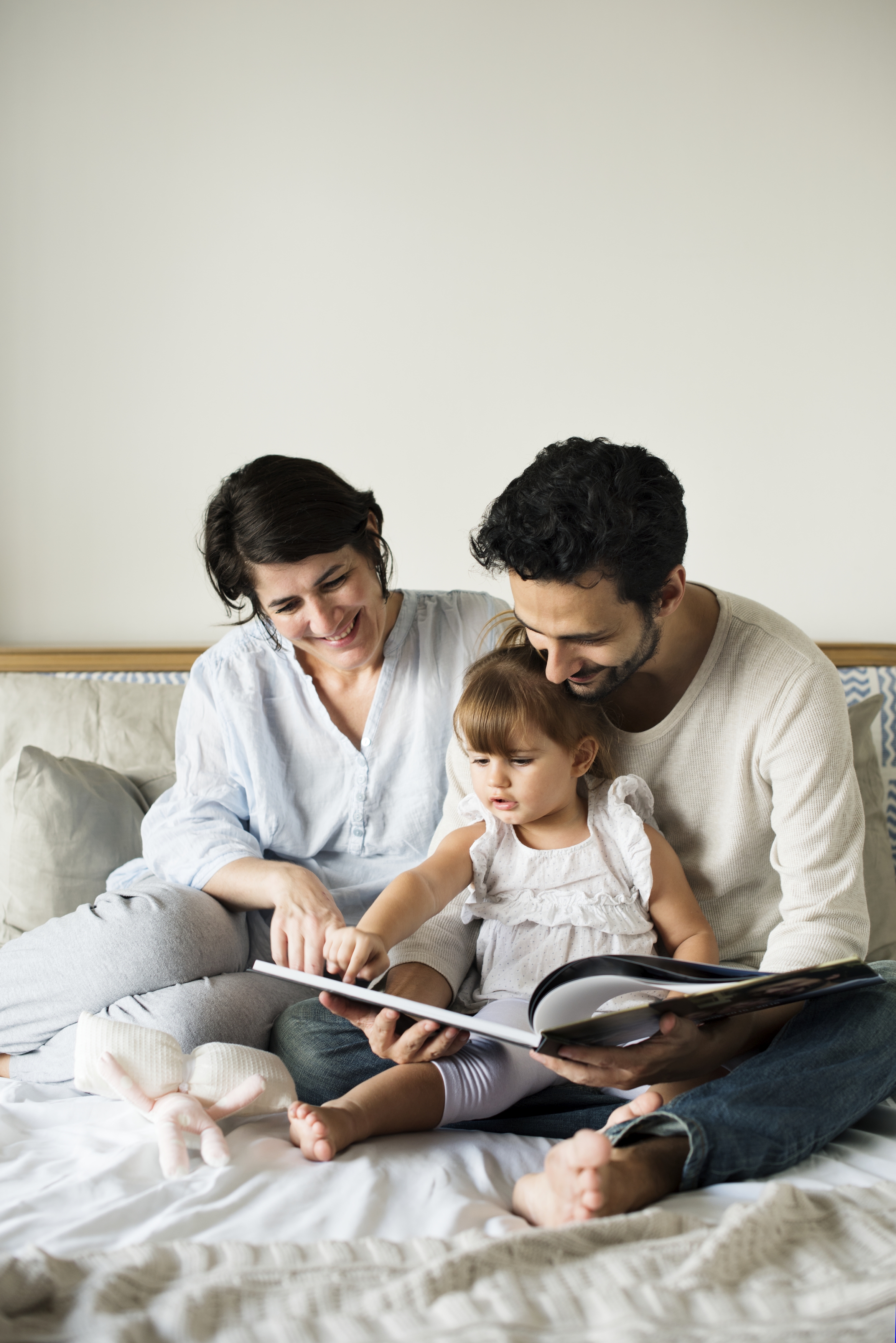 woman, man and young daughter reading a picture book together on a bed.