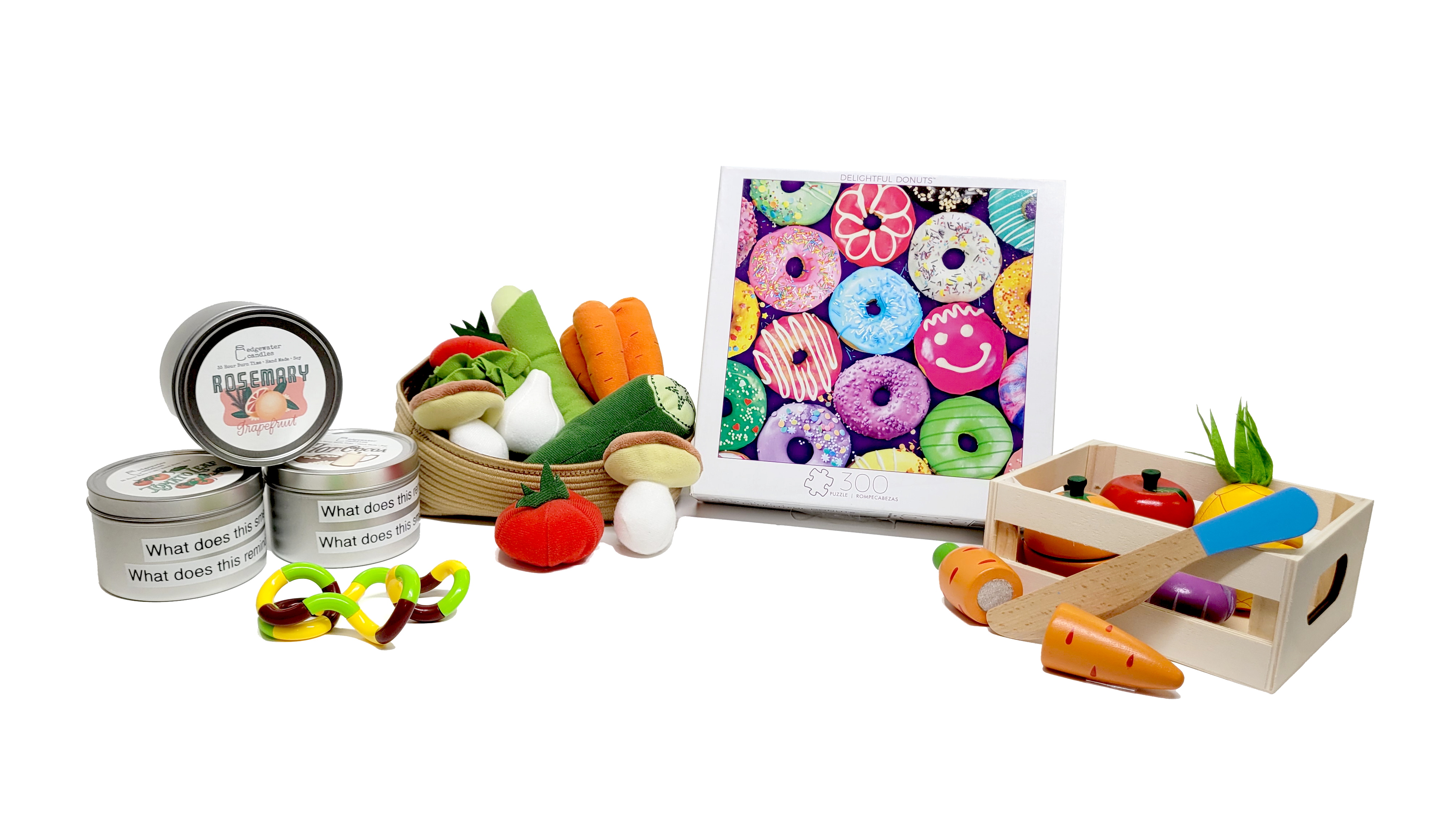 Arrangement of food and outdoors-themed items found in the memory kit, including a colorful donut puzzle, a crate of wooden fruits and vegetables, a basket of cloth vegetables, a yellow and green fidget toy, and three scented candle tins.