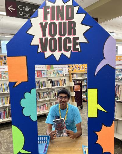 Teen summer reading volunteer sitting at the summer reading booth holding up a book 