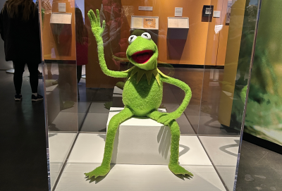Kermit the Frog sitting in a museum display case waving 