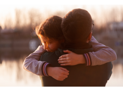an adult hugging a child with a blurred lake in the background.